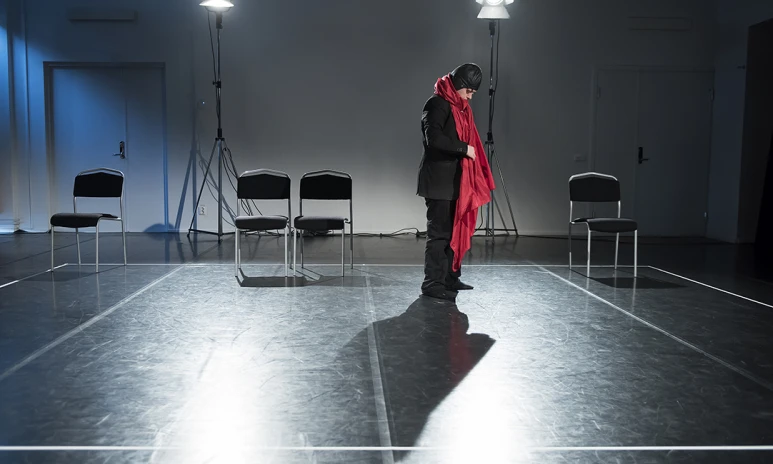 En person i svarta kläder och mössa med stor röd halsduk står ensam på en scen och blickar ned.