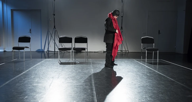 En person i svarta kläder och mössa med stor röd halsduk står ensam på en scen och blickar ned.