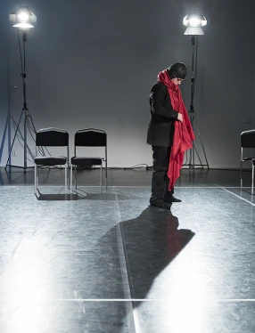 En person i svarta kläder och mössa med stor röd halsduk står ensam på en scen och blickar ned.