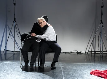 två personer sitter bredvid varandra på två stolar på scenen och kramar om varandra.