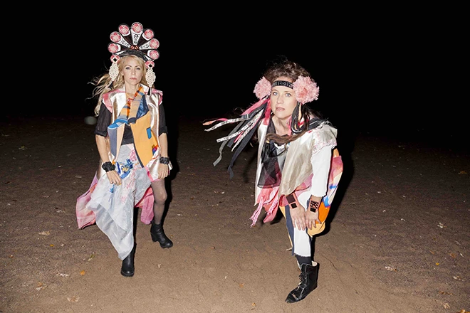 Maria och Cecilia ute i mörkret. De står i sanden och har färglada folkloristiskt insprirerade dräkter och huvudbonader på sig