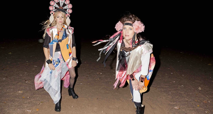Maria och Cecilia ute i mörkret. De står i sanden och har färglada folkloristiskt insprirerade dräkter och huvudbonader på sig
