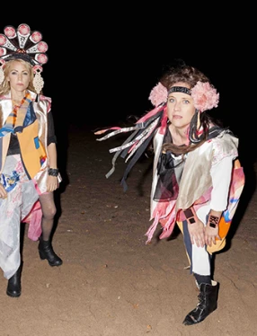 Maria och Cecilia ute i mörkret. De står i sanden och har färglada folkloristiskt insprirerade dräkter och huvudbonader på sig