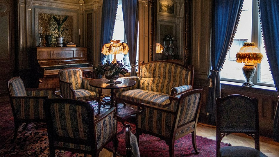 Salong med sittmöbler och piano, stora lampor och tunga gardiner.
