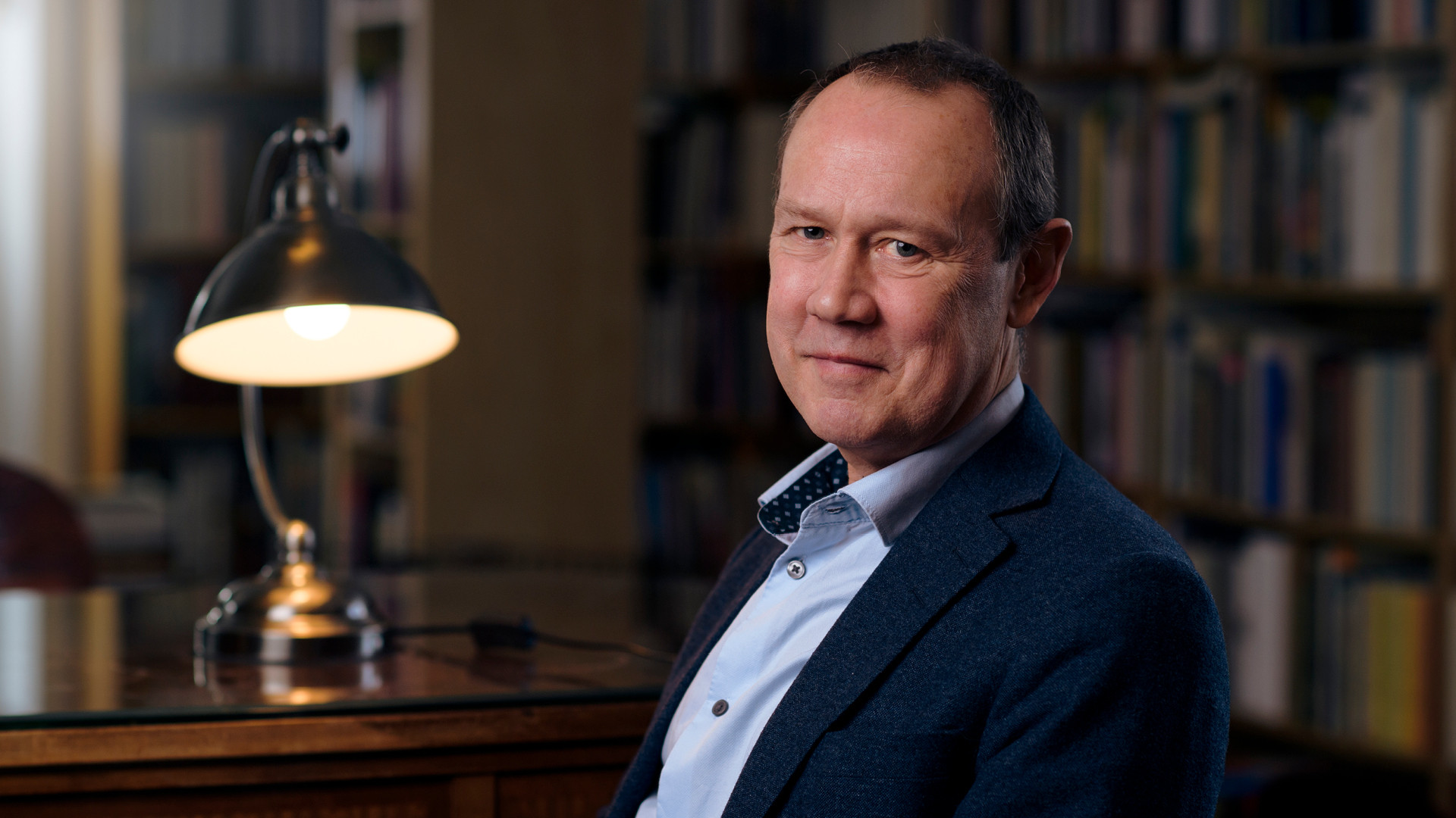 Foto av Bo Eriksson sittande i ett bibliotek med en lampa bredvid.