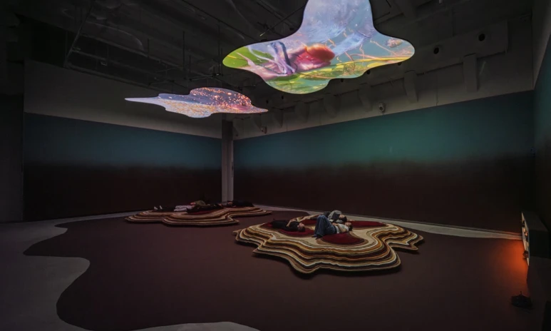 Övergripande vy av två asymmetrisk former i taket där två videofilmer projiceras, personer ligger under dessa på två högar asymmetriska mattor.
