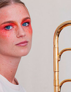 Blond kvinna med blå ögon och orange ögonskugga runt ögonen. Håller i en trombon. Foto.