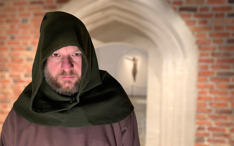 Man i medeltida kläder vid uppbyggd miljö av medeltida tegelstenskyrka.